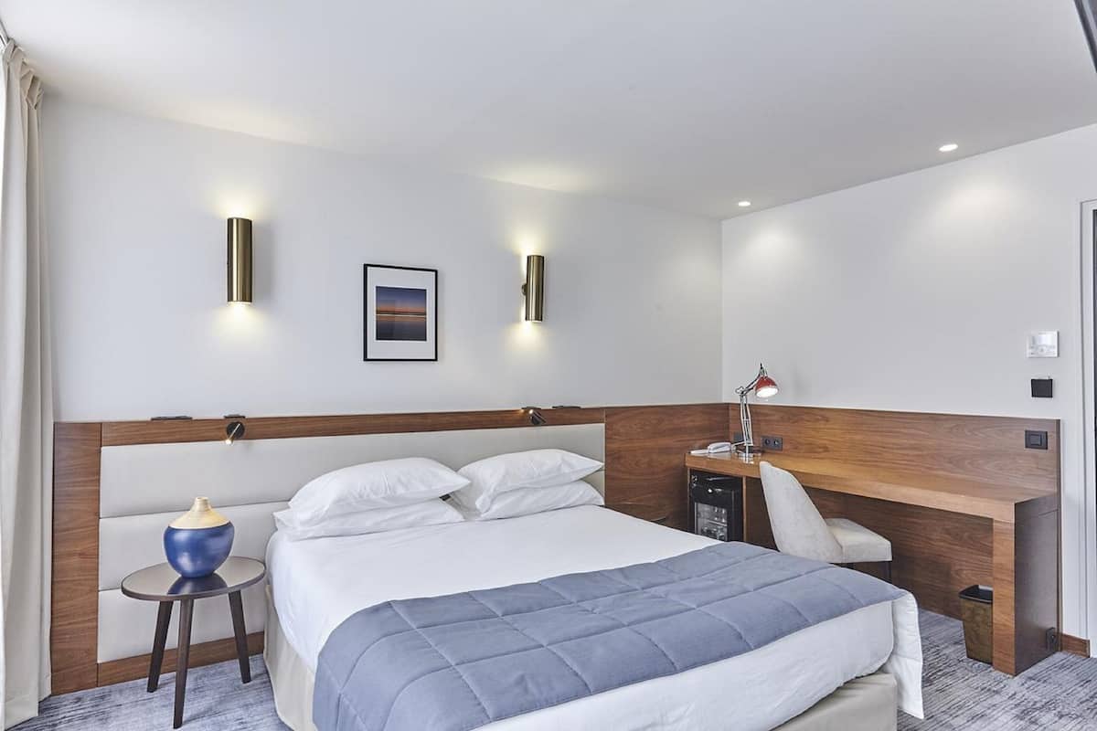 Una habitación de hotel moderna con una cama grande, una mesita de noche con lámpara, una pequeña mesa auxiliar con un florero y un escritorio de madera con una silla.