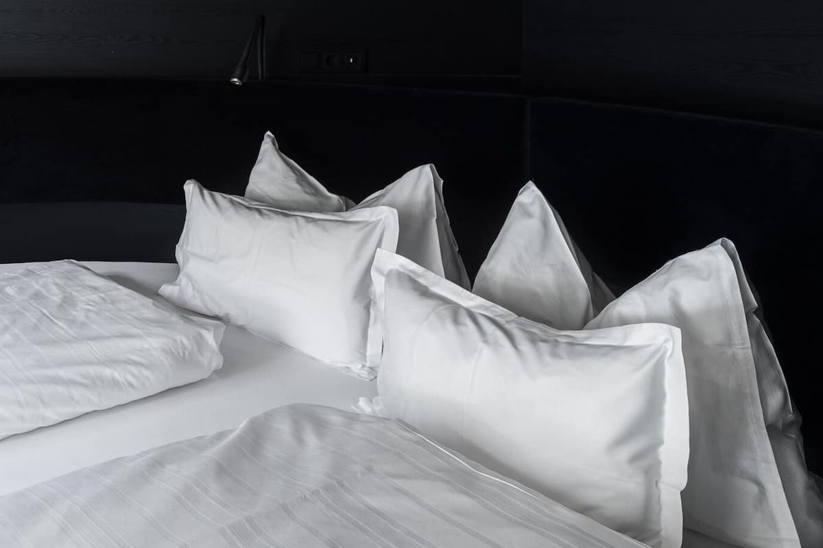 Un letto con lenzuola bianche e numerosi cuscini, una testiera scura e una parete con una texture in legno.