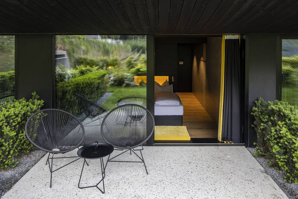 Zona esterna moderna con posti a sedere, due sedie e un tavolino, che conduce a una camera da letto con un letto e un tappeto giallo.