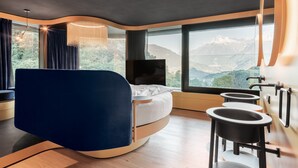 A modern hotel room with a curved bed, a TV, a small bathroom, and a large window offering a mountain view. - Prinz Rudolf Smarthotel (Merano)