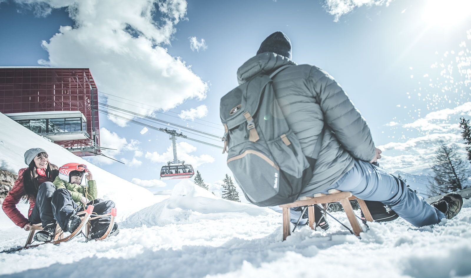 Eine Person sitzt auf einem hölzernen Stuhl in verschneiter Landschaft, im Hintergrund ist eine Seilbahn zu sehen.