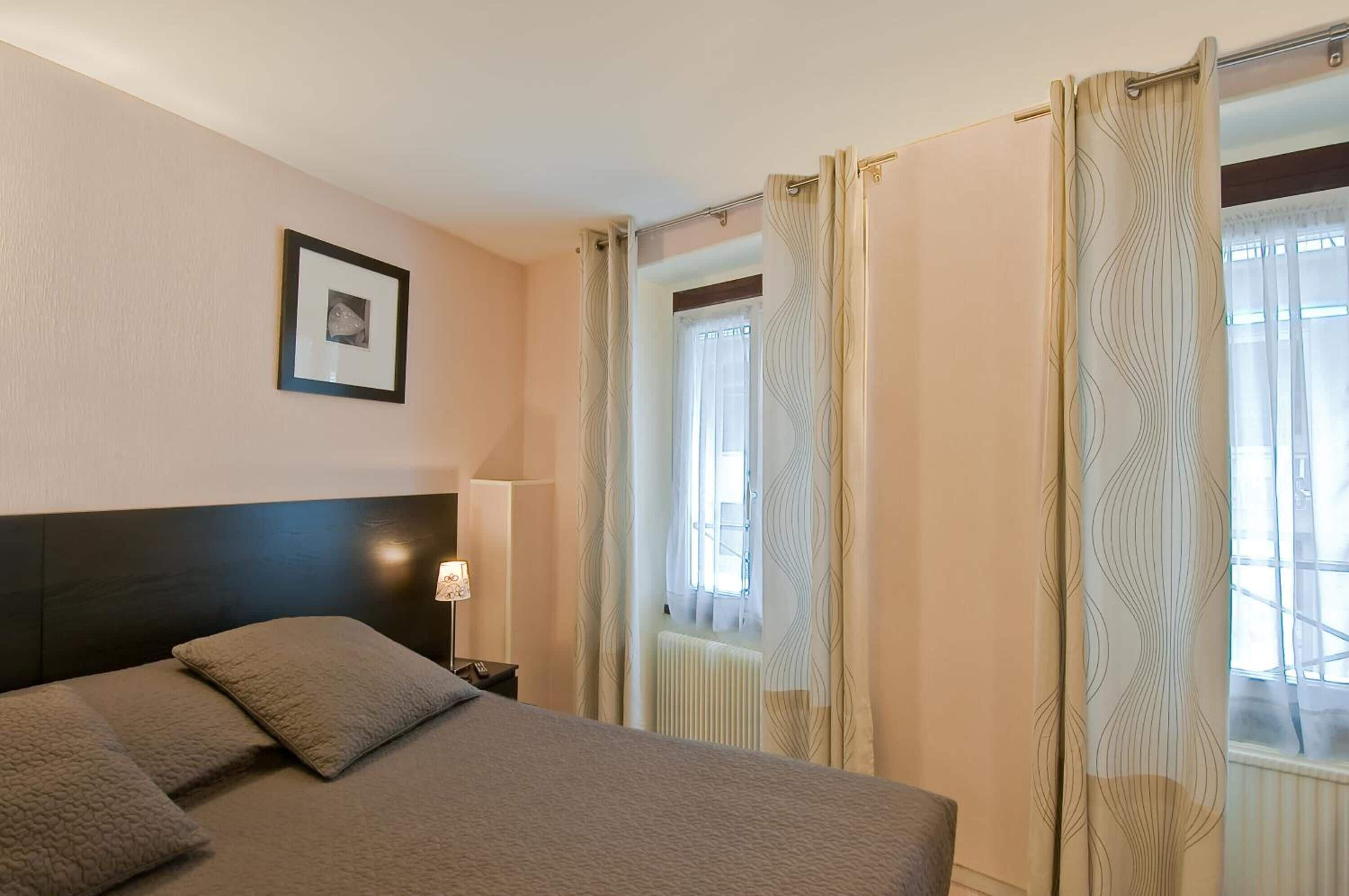 Une chambre d’hôtel avec un lit, une table de chevet, une lampe, un tableau encadré au mur et des rideaux aux fenêtres.