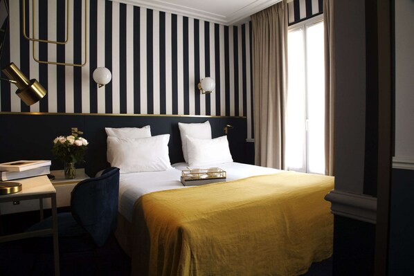 Habitación de hotel con pared a rayas, una cama con ropa blanca, una mesita de noche con lámpara y una ventana con cortinas.