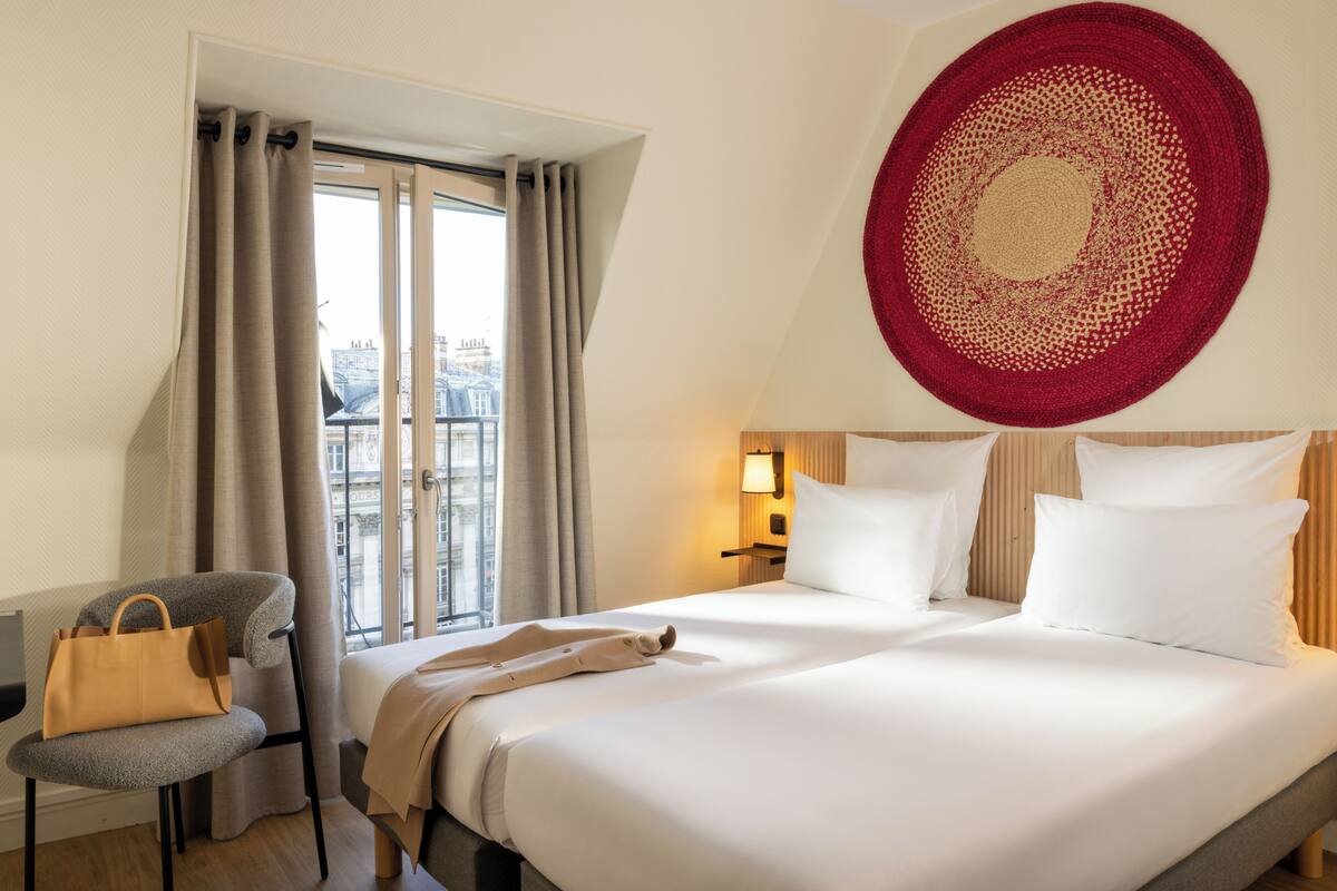 A hotel room with a bed, a chair, a handbag, a window with curtains, and a wall-mounted circular rug.