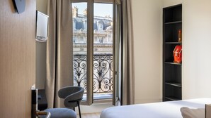 Une chambre d’hôtel avec un lit, une chaise, une télévision, une bibliothèque et un balcon donnant sur des immeubles.