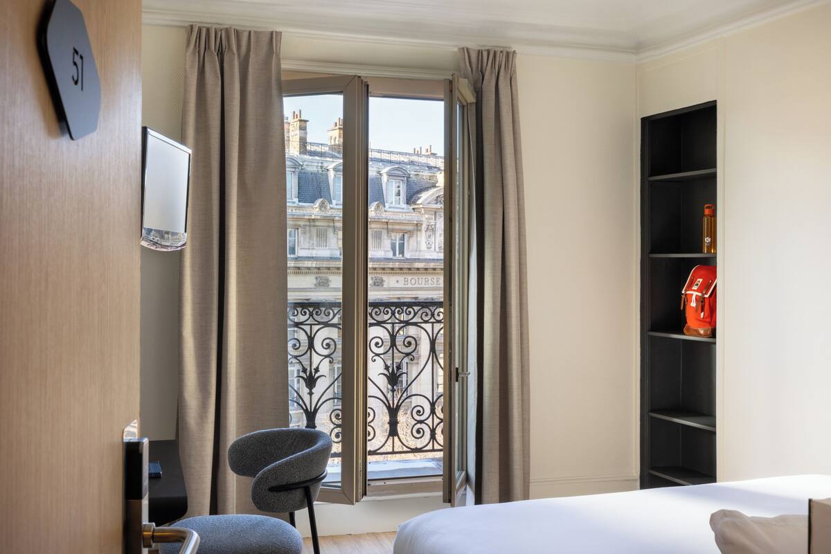 A hotel room with a bed, a chair, a TV, a bookshelf, and a balcony with a view of buildings.