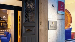 L’entrée d’un hôtel, avec une porte sombre, une enseigne indiquant « Korner République », et une vitrine où l’on aperçoit une chaise bleue et une plante en pot.