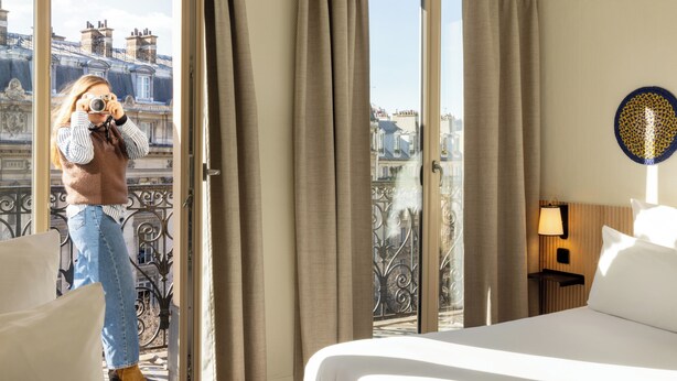 Une chambre d’hôtel avec un lit, une table de chevet avec une lampe et une vue sur la ville grâce à une grande fenêtre dotée de rideaux.