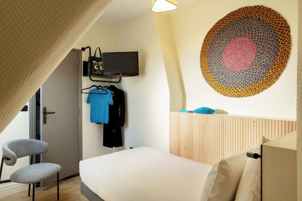 A bedroom with a bed, a TV mounted on the wall, a chair, and a coat rack with clothes.