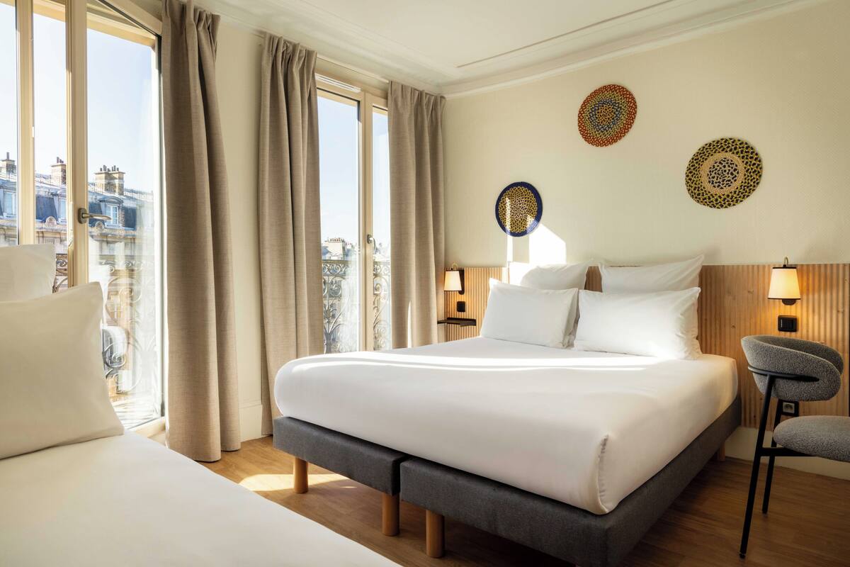 A hotel room with a large bed, a bench, two bedside tables with lamps, and a view of the city.