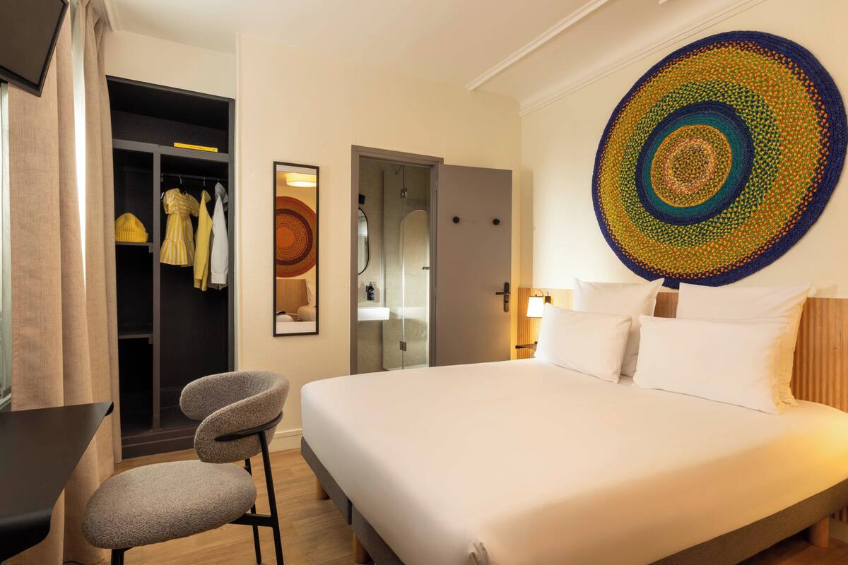 A hotel room with a bed, a chair, a wardrobe, and a large circular artwork on the wall.