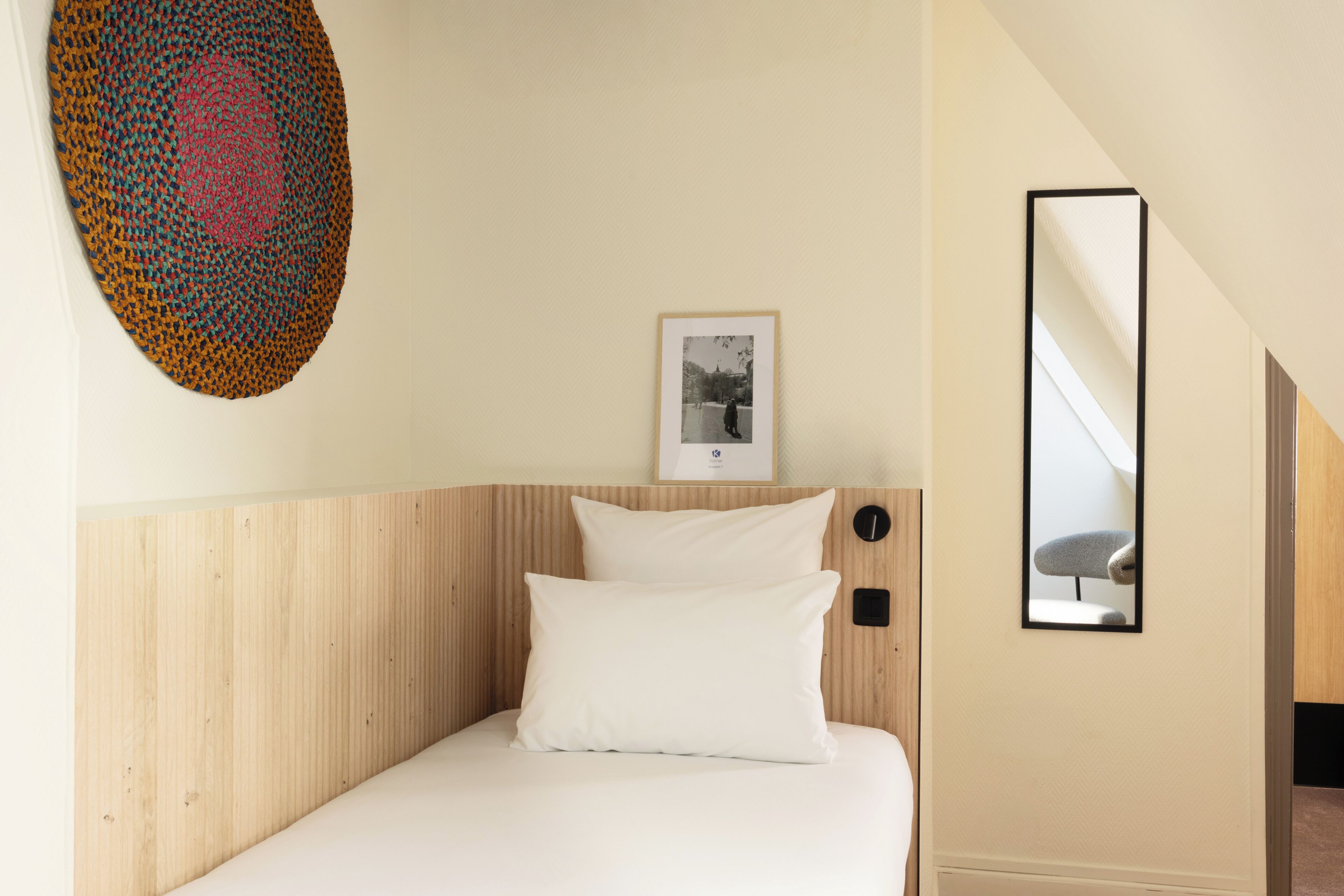 A hotel room with a wooden headboard, a bed with white bedding, a framed photograph on the wall, and a mirror reflecting part of the room.