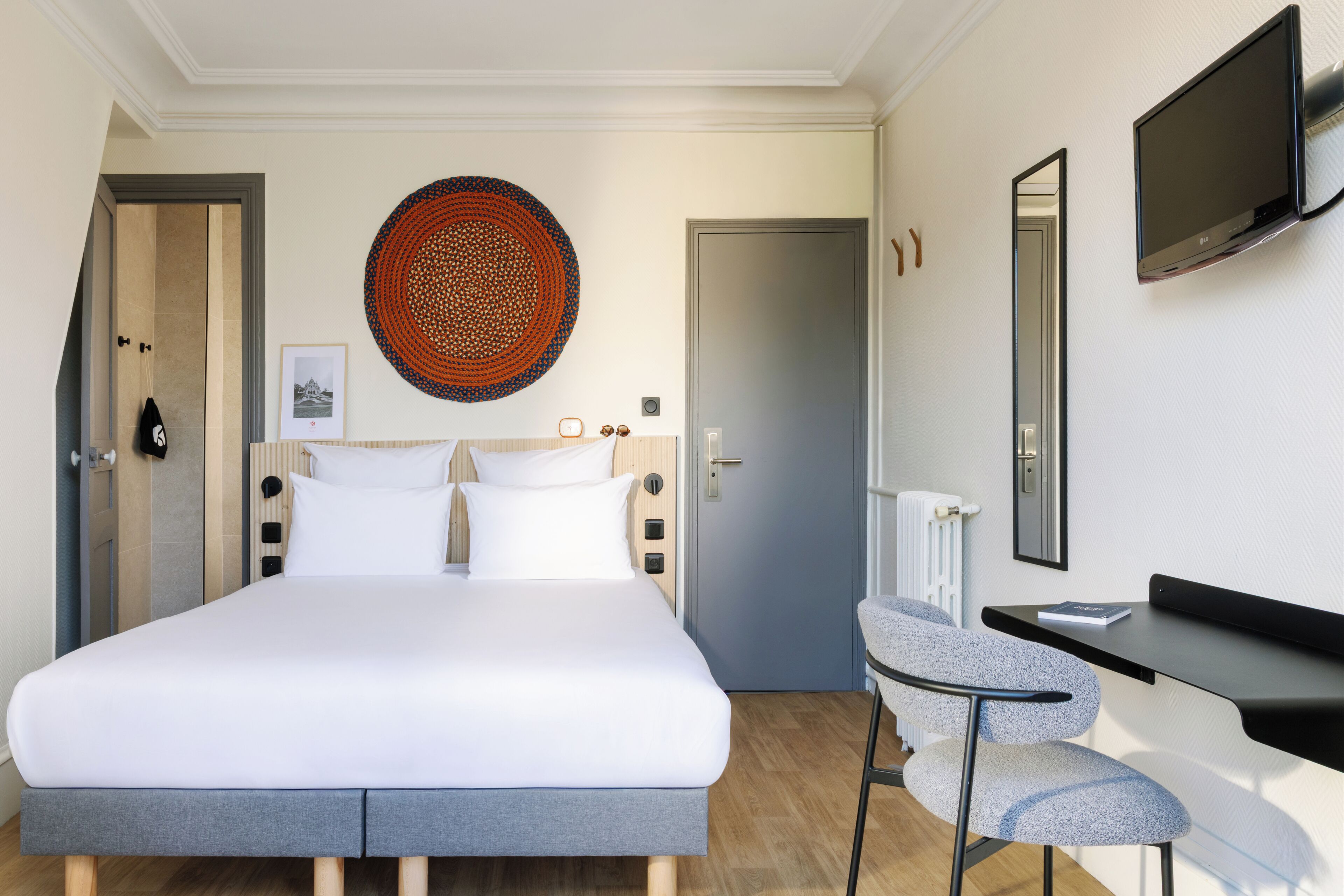 A hotel room with a bed, a desk, a chair, a television, a door, and a wall decoration.