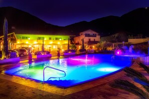 Ein Schwimmbecken mit farbigem Licht, ein überdachter Bereich mit Sonnenschirmen und ein Gebäude mit zahlreichen Fenstern und Lichtern.