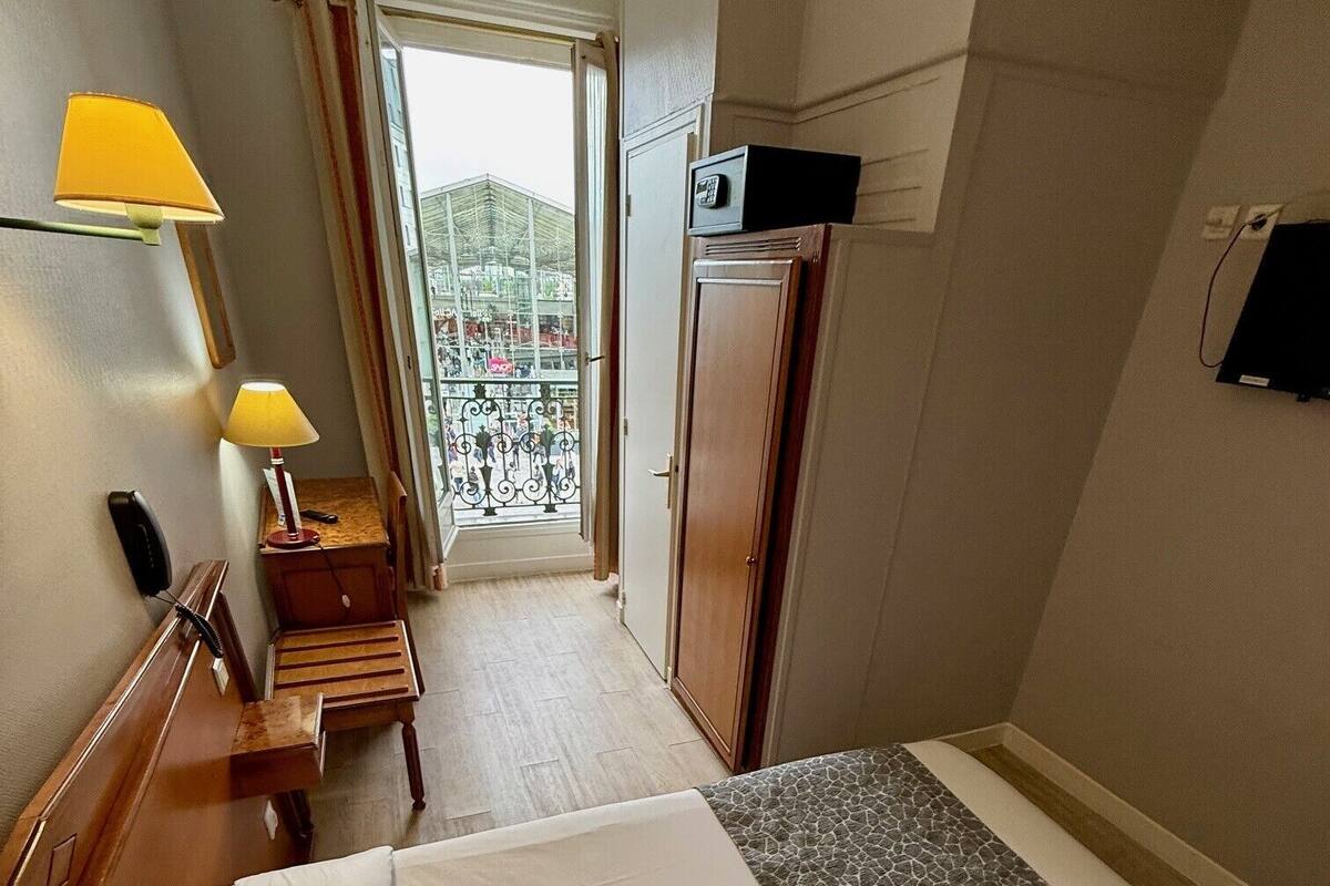 A hotel room with a bed, a desk with a lamp, a wardrobe, and a view of the outside balcony.