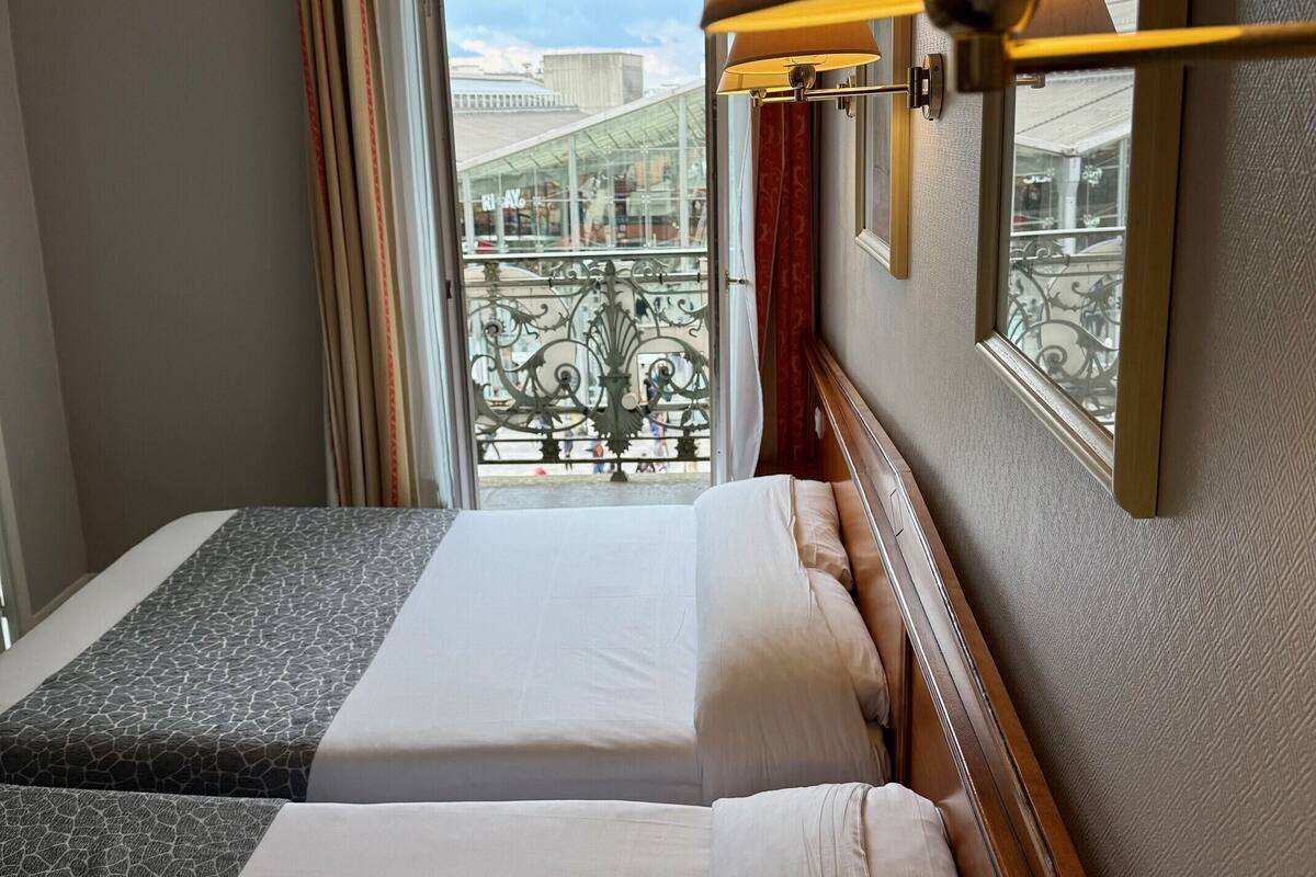 A hotel room with two beds, a balcony with a view of the city, and a lamp on the wall.