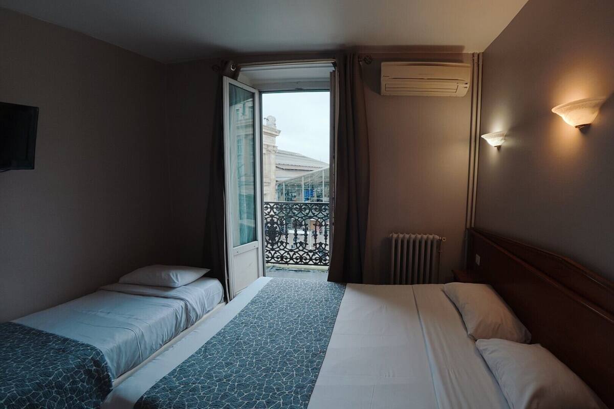 A hotel room with two single beds, a large window with a view of buildings, an air conditioning unit, and a wall-mounted light fixture.