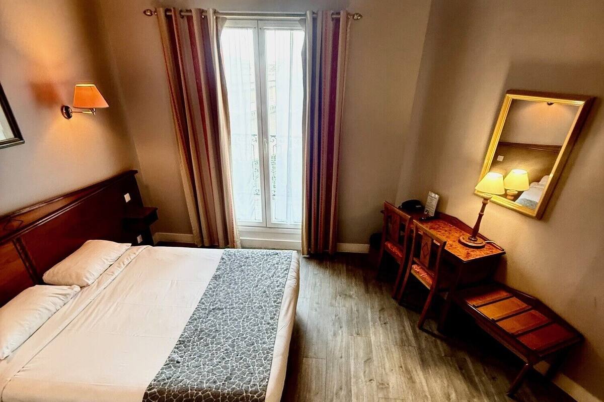 A hotel room with a bed, a desk, a chair, a mirror, and a window with curtains.