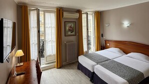 Una habitación de hotel con cama, escritorio, silla, lámpara y ventana con cortinas.