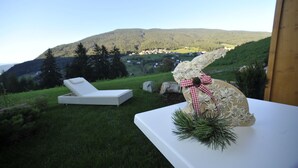 A white lounge chair and a decorative bunny on a table with a view of a hillside and a village. - Hotel Digon (Castelrotto)