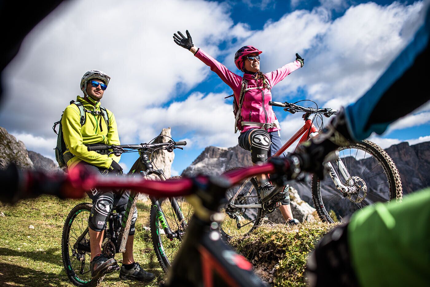Tres ciclistas de montaña en un sendero cubierto de hierba, con montañas rocosas al fondo.
