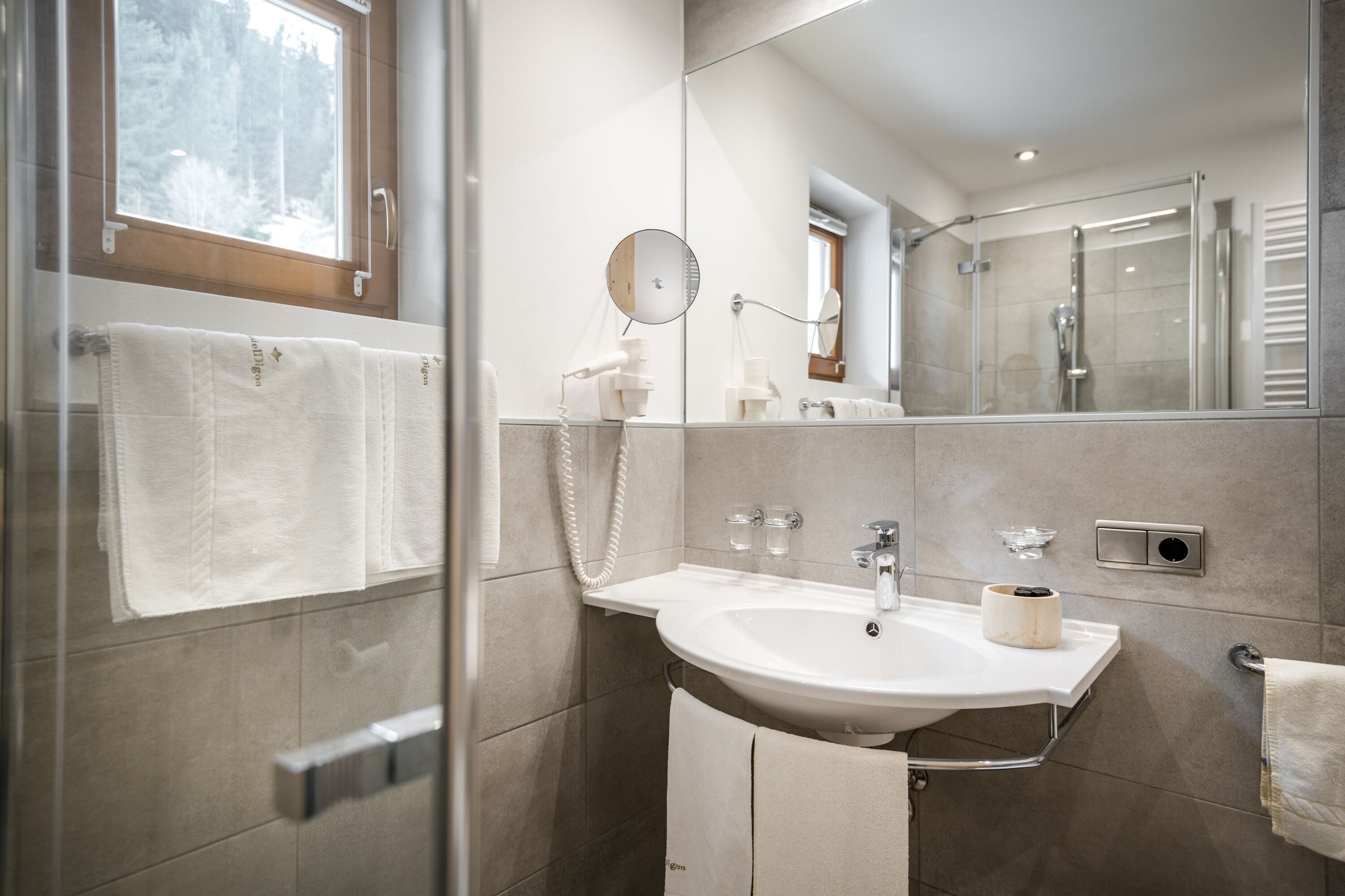 A modern bathroom with a glass shower enclosure, a large mirror, a sink with a towel rack, and a window with a view of trees.