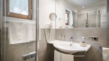 A modern bathroom with a glass shower enclosure, a large mirror, a sink with a towel rack, and a window with a view of trees.