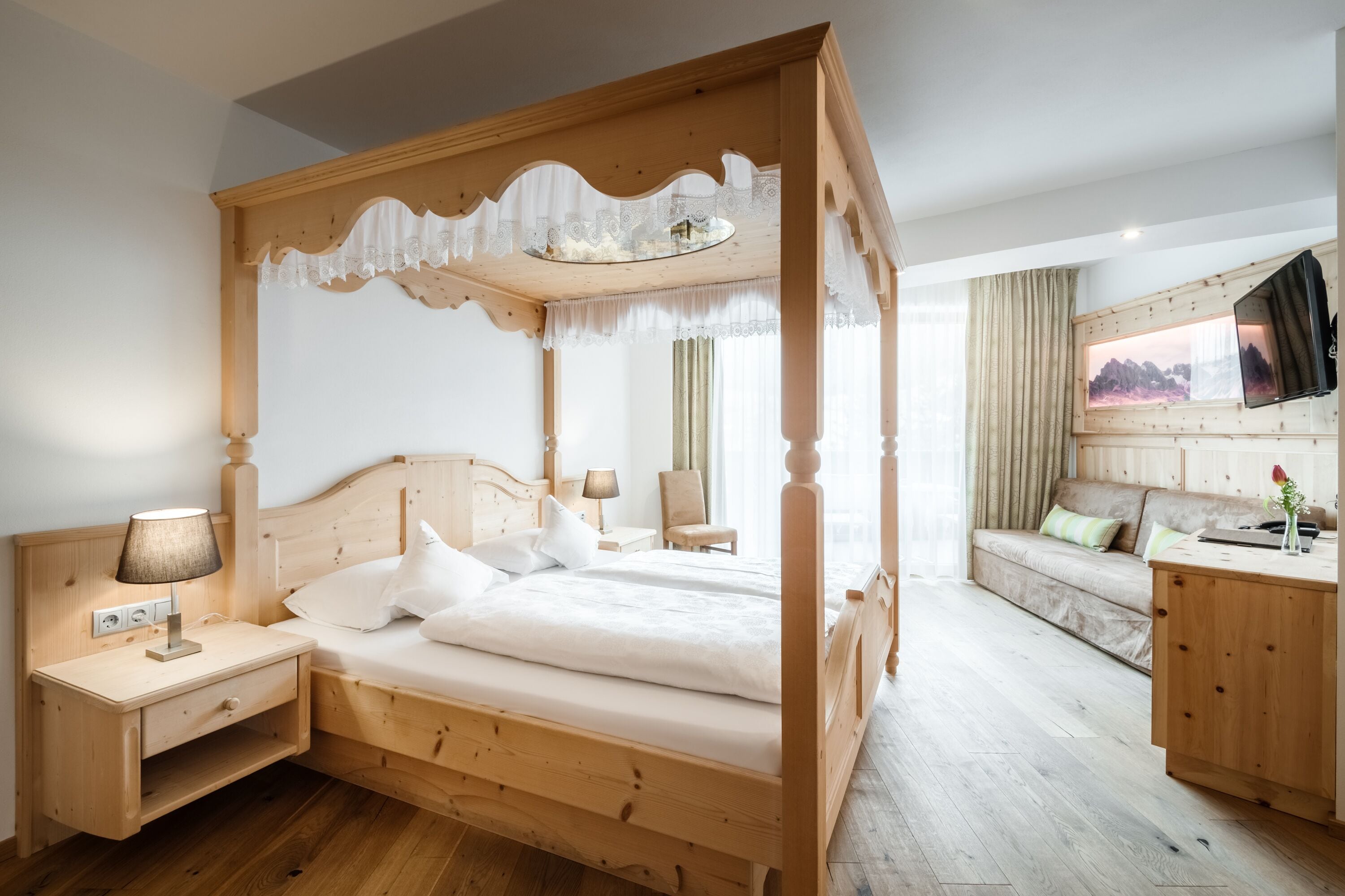 Una cama con dosel de madera con ropa de cama blanca, una mesita de noche con lámpara, un tocador de madera, un sofá y un escritorio con espejo.