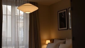 A hotel room with a bed, a bedside lamp, a window with sheer curtains, and a hanging light fixture.