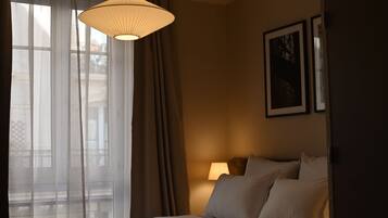 A hotel room with a bed, a bedside lamp, a window with sheer curtains, and a hanging light fixture.