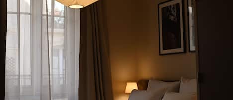 A hotel room with a bed, a bedside lamp, a window with sheer curtains, and a hanging light fixture.