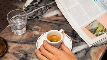 A hand holding a cup of coffee on a marble table with a newspaper and a glass of water.