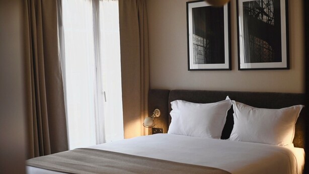 A hotel room with a bed, curtains, a nightstand, a wall-mounted lamp, and two framed pictures. - Local 101 Beaurepaire (Paris)
