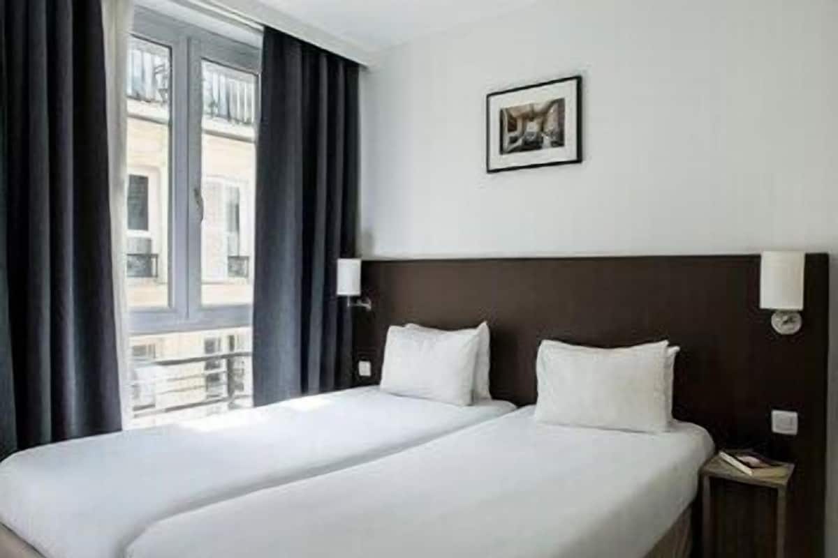 A hotel room with two beds, a window with curtains, a framed picture on the wall, and a nightstand with a lamp.