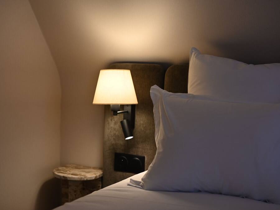 A hotel room with a bed, pillows, a nightstand with a lamp, and a small round table.