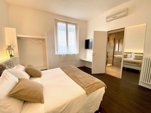 Un dormitorio con cama, mesita de noche, televisor, armario y baño visible a través de una puerta abierta.