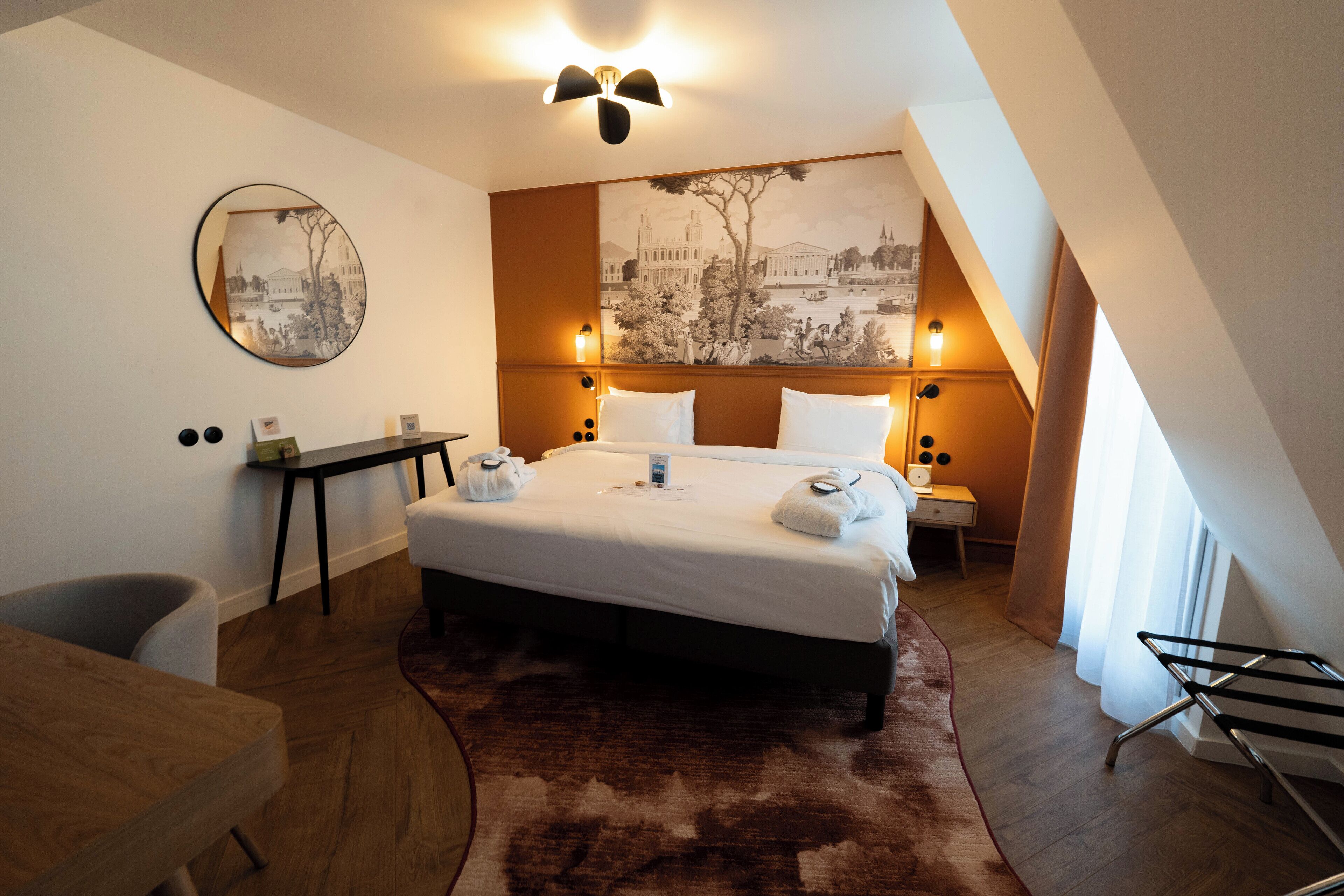 Habitación de hotel con una cama grande, un escritorio, una silla, un espejo redondo y una pared con un cuadro de un paisaje urbano.