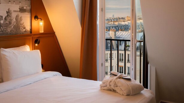 A hotel room with a bed, a nightstand, a wall-mounted lamp, a window with a view of buildings, and a painting on the wall.