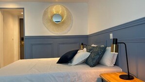 Una cama bien hecha con edredón blanco, un espejo redondo en la pared y una mesita de noche con lámpara.