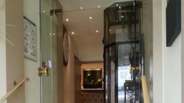 Entrada de un hotel con puerta de cristal, una escalera que lleva al vestíbulo y un pequeño jardín interior visible a través del vidrio.