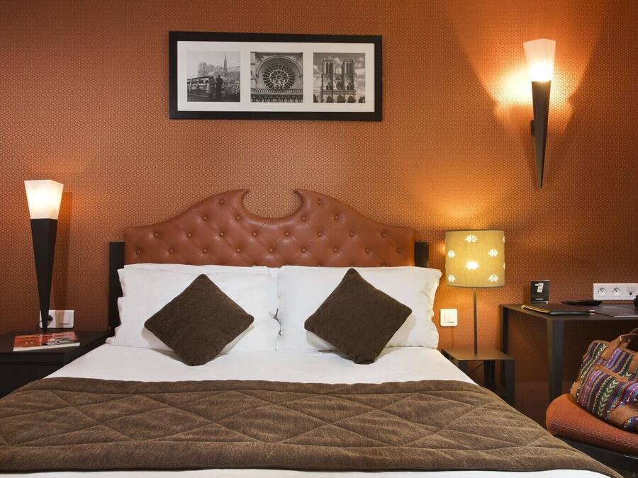 A hotel room with a tufted headboard, a quilted bedspread, two pillows, a framed picture above the bed, a nightstand with a lamp, and a chair with a handbag.