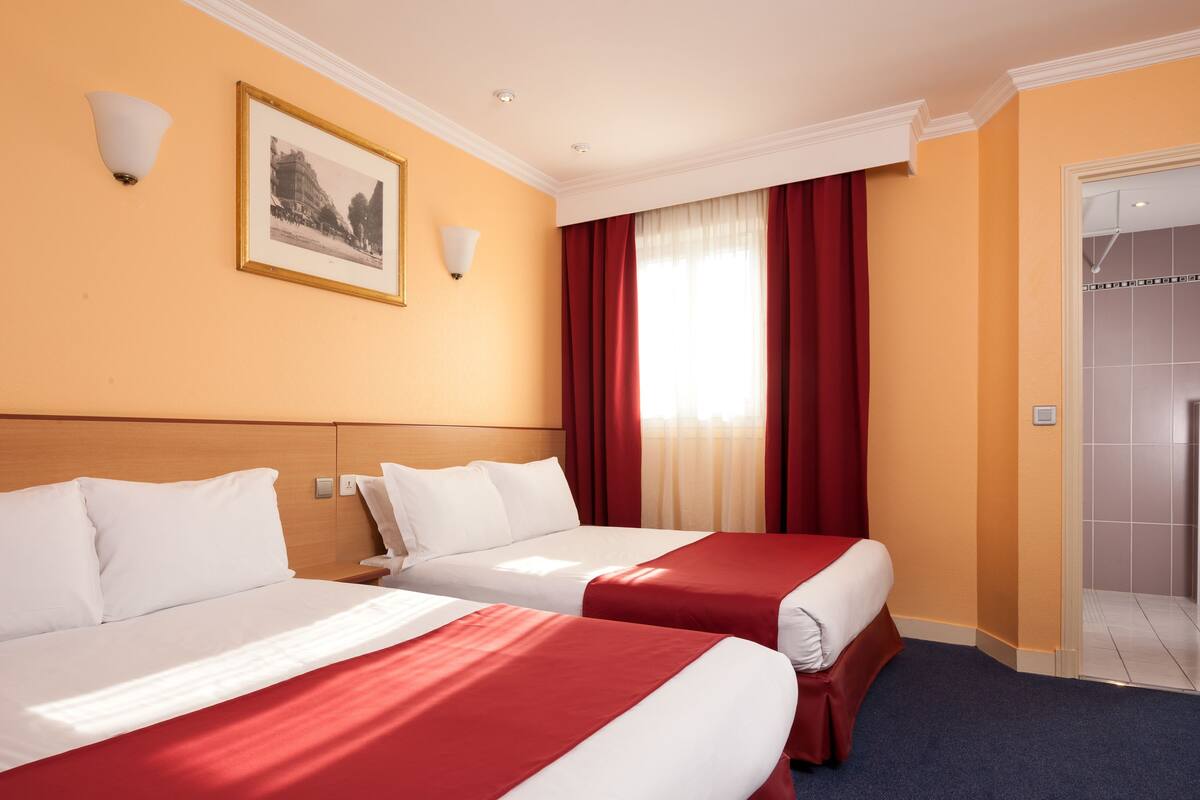 A hotel room with two beds, a framed picture on the wall, a window with red curtains, and a shower area.