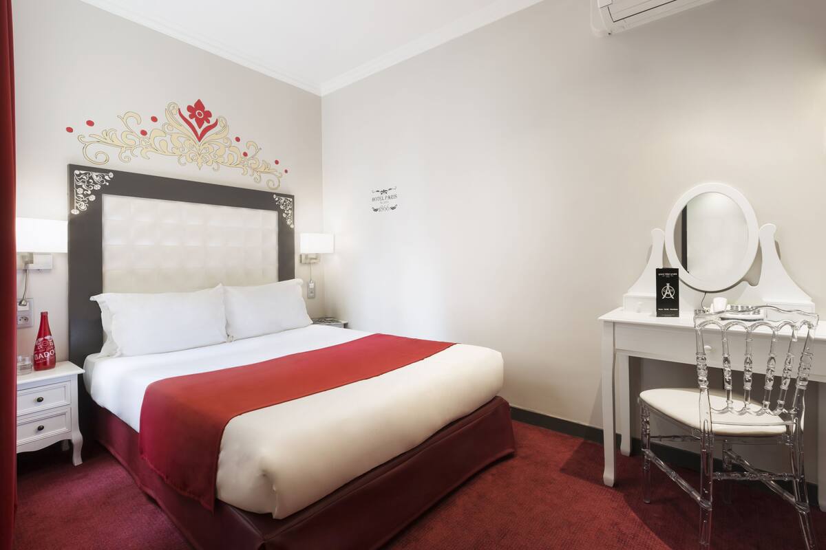 A hotel room with a bed, a desk with a mirror, a nightstand, and a decorative wall piece.