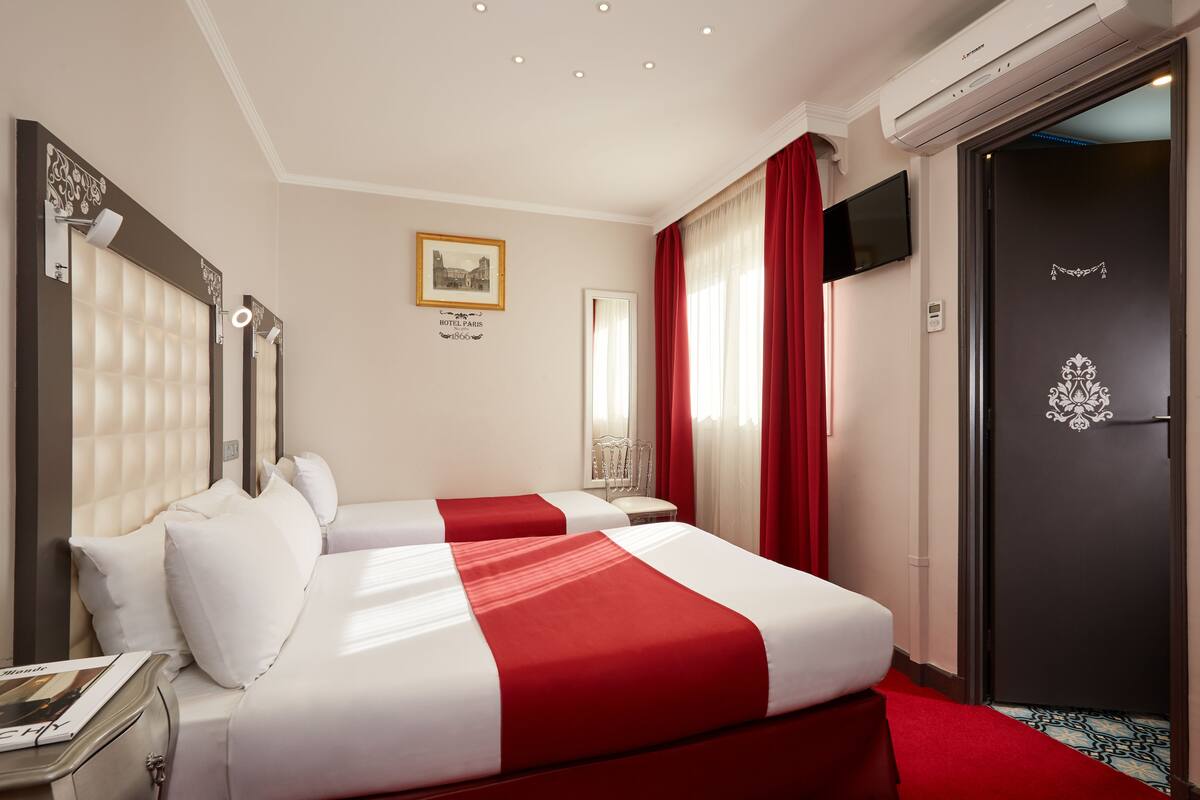 A hotel room with two beds, a red and white bedspread, a framed picture on the wall, and a door with a decorative emblem.