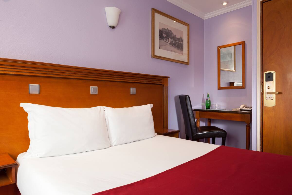 A hotel room with a wooden headboard, a bed with white linens, a red throw blanket, a desk with a telephone, a chair, and a framed picture on the wall.