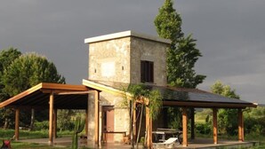 Un edificio de piedra con techo plano, un espacio al aire libre cubierto con una mesa y sillas, y vistas a la vegetación circundante.