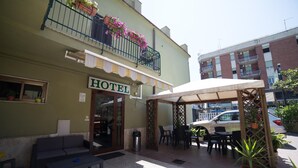 A hotel with a covered outdoor seating area, a balcony with flowers, and a sign indicating the hotel's name. - Le Rose (Tivoli)