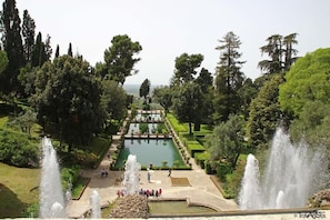 Um jardim formal com uma fonte central, sebes bem aparadas e diversas árvores.