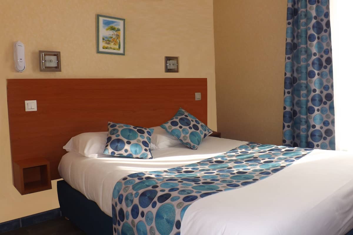 Una cama bien hecha con colcha azul y blanca estampada, dos almohadas con un diseño similar, un cabecero de madera, un pequeño cuadro en la pared y una ventana con cortinas azules.