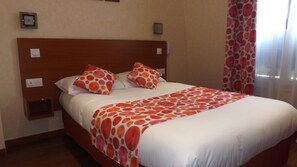 Una habitación de hotel con cabecera de madera, una cama con ropa blanca y almohadas de estampado naranja, una mesita de noche con lámpara y una ventana con cortinas translúcidas.